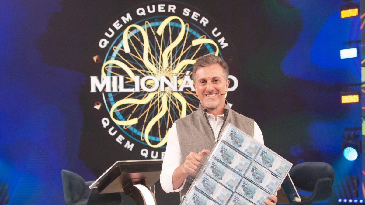 Apresentador Luciano Huck à frente de telão com banner do quadro "Quem quer ser um milionário"